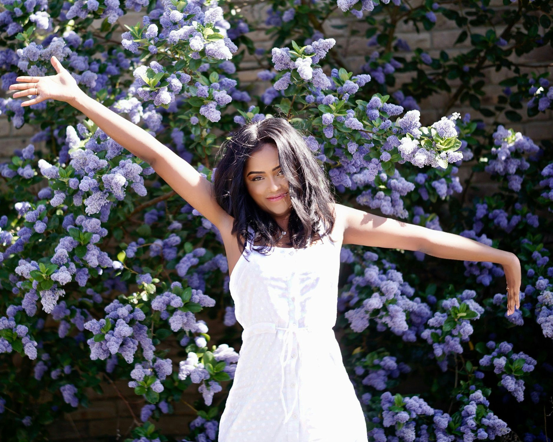 Sachini wearing a white dress in front of a tree with purple flowers